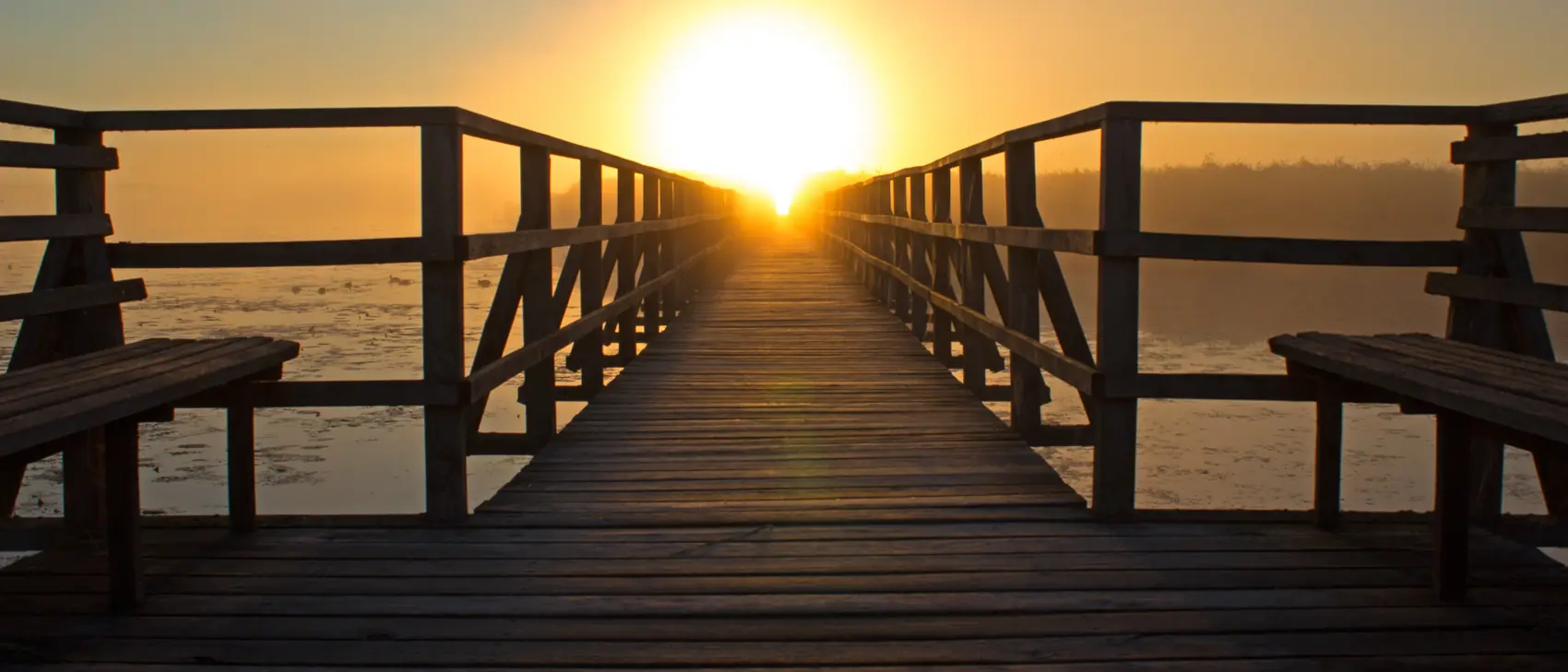 A wooden walkway over a lake pointing towards the rising sun