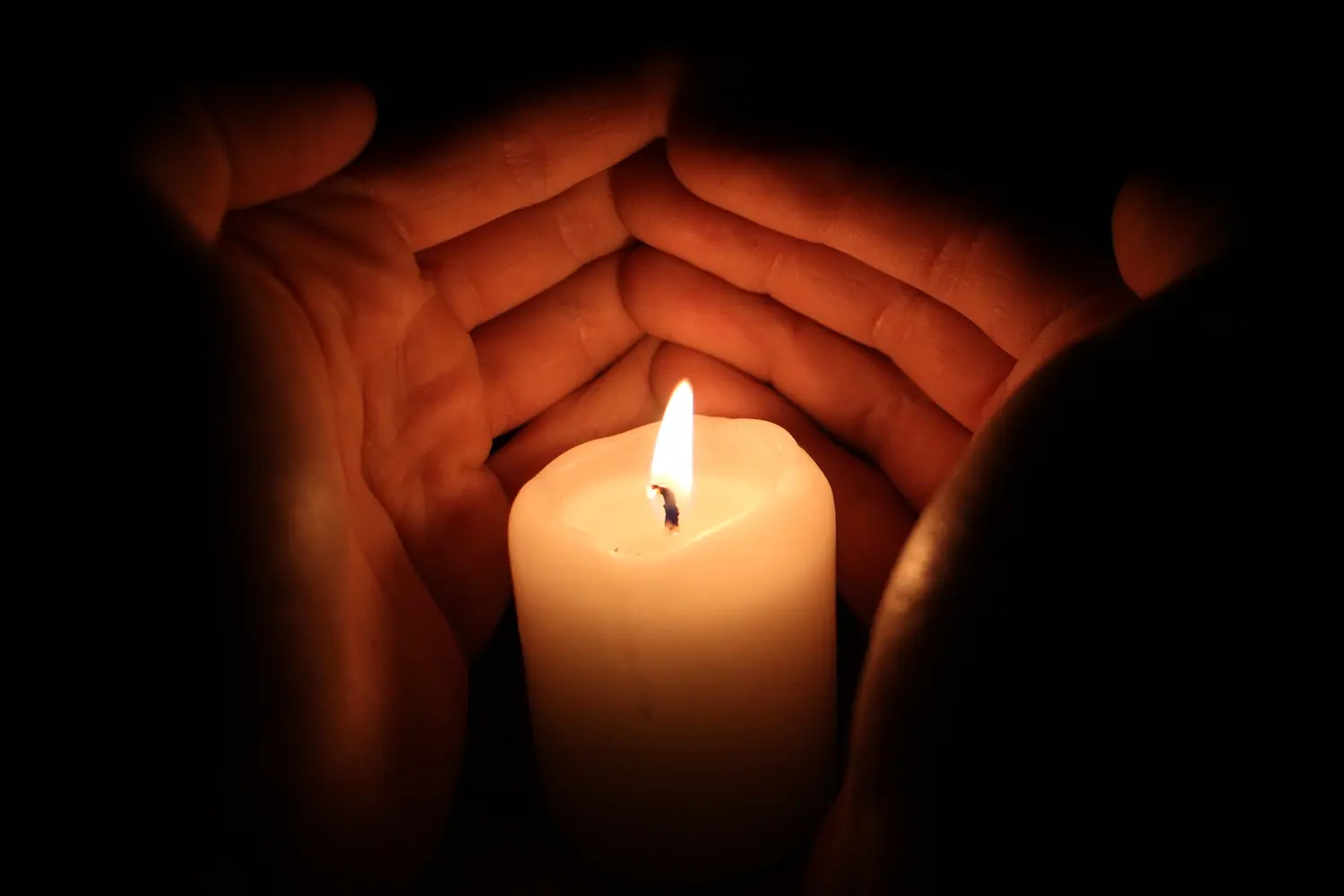 Two hands sheltering the flame of a lit candle in the dark